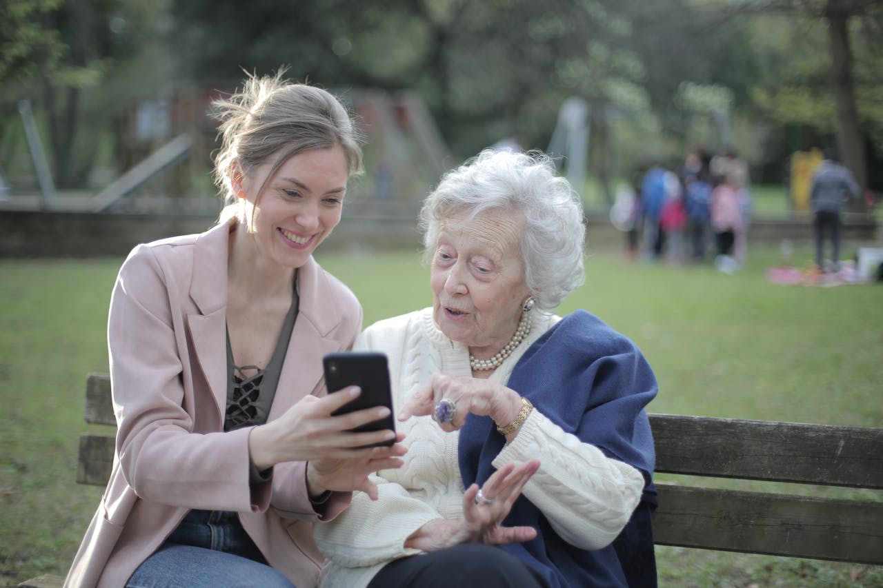 services-01 Delighted female relatives sitting together on wooden bench in park and browsing mobile phone while learning using